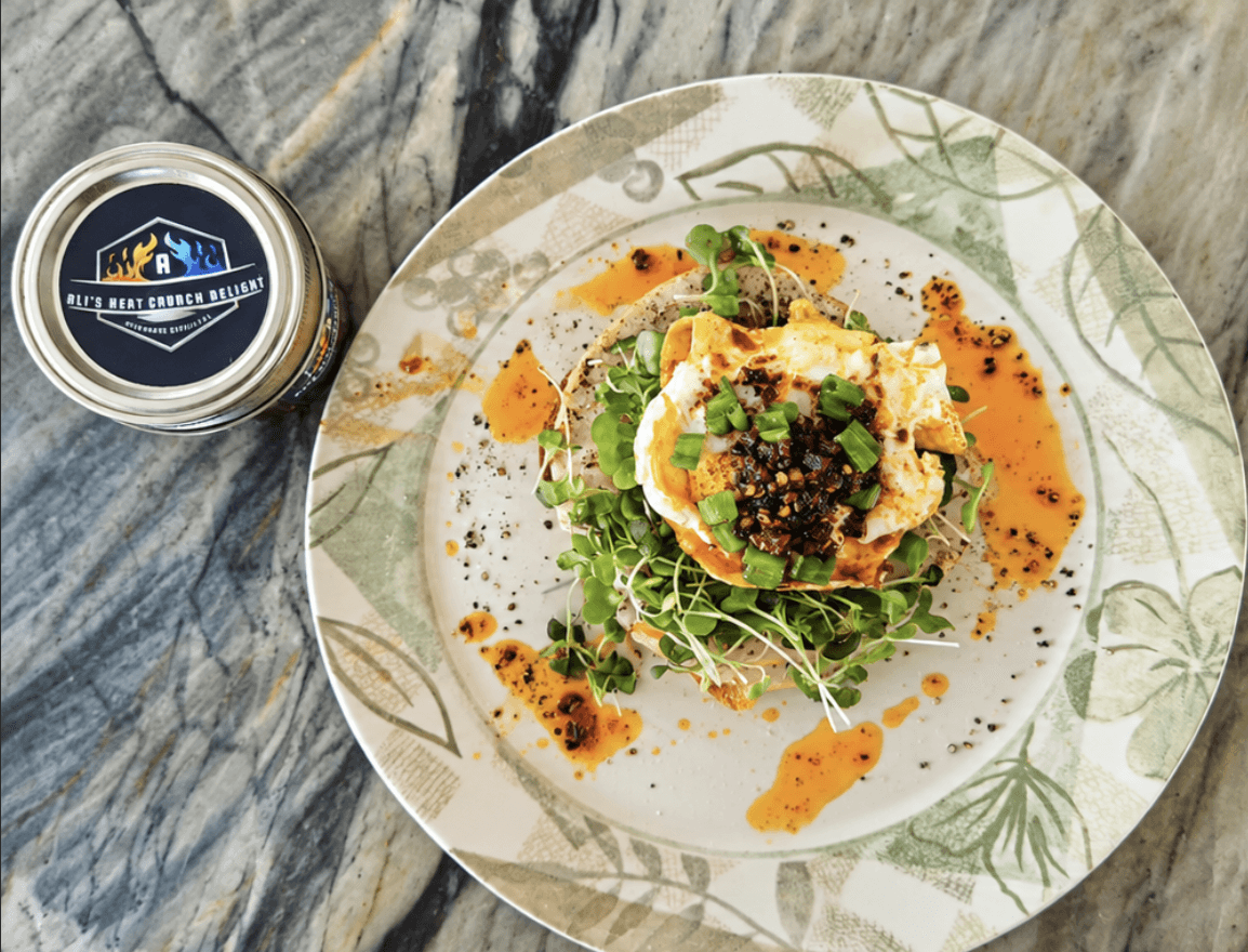 A plate of toast topped with microgreens, a fried egg and a spoonful of Ali's Heat Crunch Delight, with a jar of the chilli oil beside the plate.