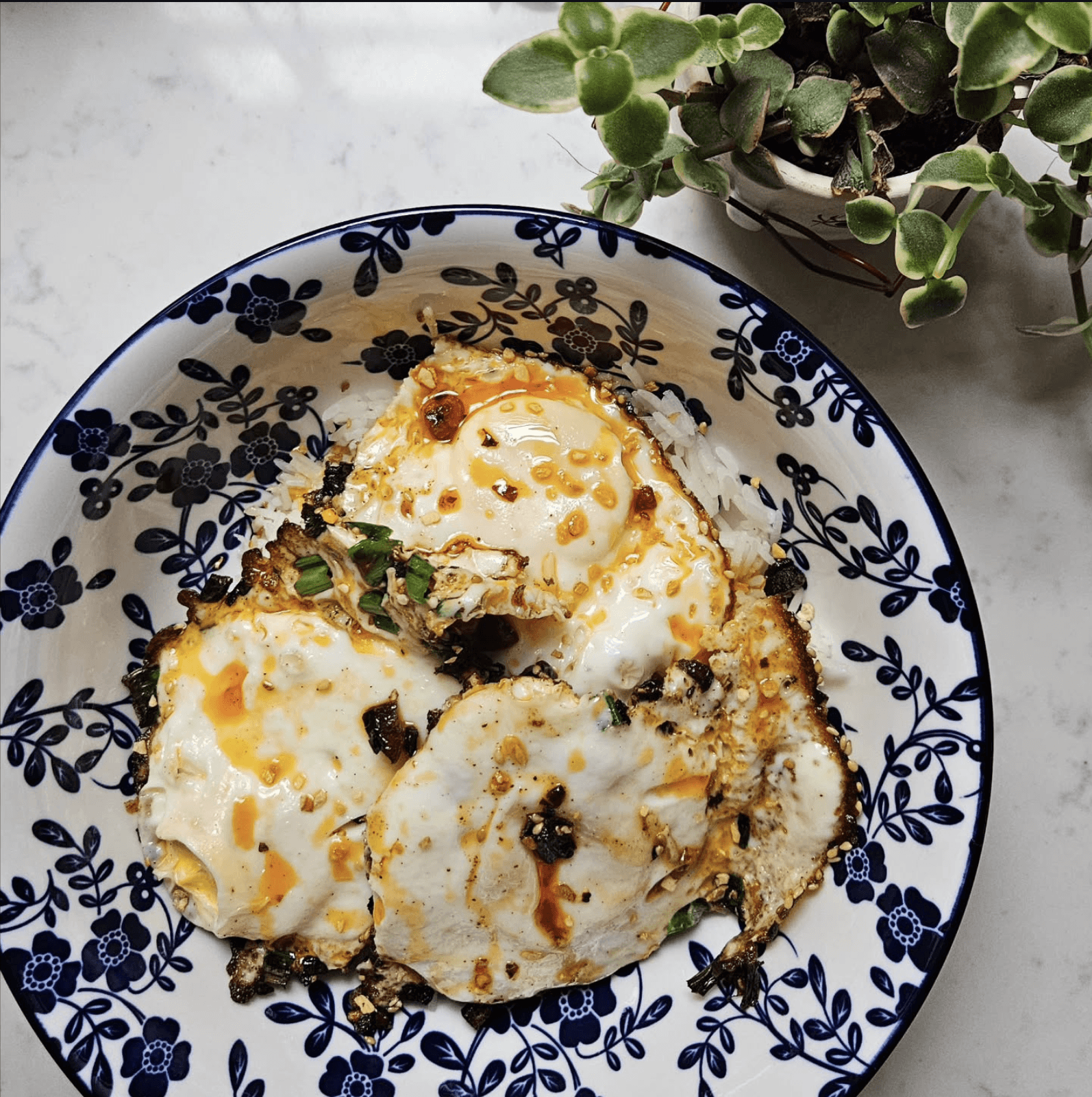 Three fried eggs over steamed rice in a blue floral bowl, drizzled with Ali's Heat Crunch Delight chilli oil.