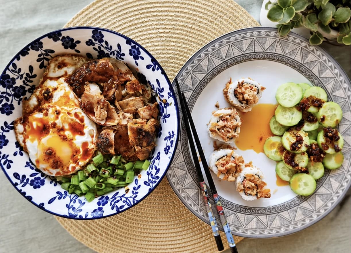 An overhead shot of two dishes: a noodle bowl topped with a fried egg, shredded chicken and chilli oil, beside a plate of sushi rolls and cucumber slices drizzled with chilli oil.
