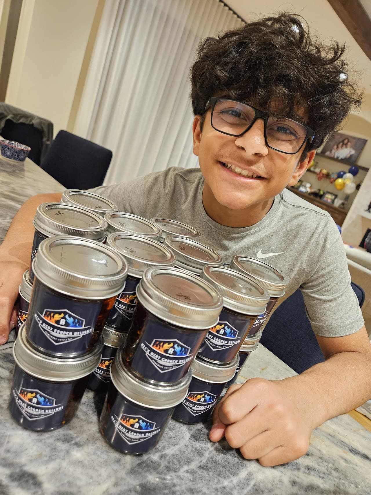 Ali smiling at a kitchen counter behind a stack of jars of Ali's Heat Crunch Delight chilli oil.